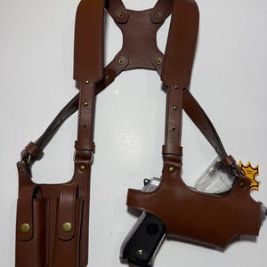 以下が含まれることがあります： 茶色のレザー製ショルダーホルスターシステム。片側にダブルマガジンポーチ、もう片側にハンドガン用ホルスターを備えています。ハーネスは調節可能なストラップ、真鍮色の金属製リベット、スナップボタン式の留め具で構成されており、装備をしっかりと収納できるよう設計されています。