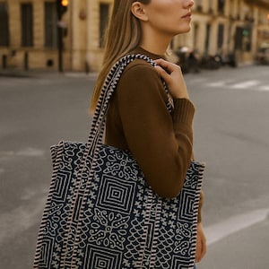 May include: A navy blue tote bag with a white and pink geometric pattern. The bag has a matching strap and trim. The person in the image is wearing a brown jumper and blue jeans.