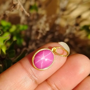 May include: A gold pendant with an oval, pink gemstone. The gemstone has a star-like pattern across its surface. The pendant is held by a finger in front of a blurred background of green foliage.