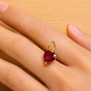 May include: A heart-shaped pendant with a deep red gemstone, set in gold-coloured metal. The pendant has a small loop at the top for a chain. The gemstone is faceted, catching the light. The pendant is a piece of jewellery.