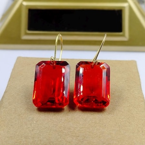 May include: A pair of red rectangular earrings with gold-colored hooks. The earrings have a faceted design, catching the light. The earrings are displayed on a tan surface.