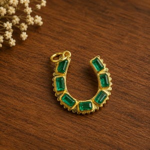 Peut inclure: Pendentif en forme de fer à cheval doré orné de pierres précieuses rectangulaires vert émeraude. Le pendentif comporte une petite boucle pour une chaîne. Sur une surface en bois brun foncé.