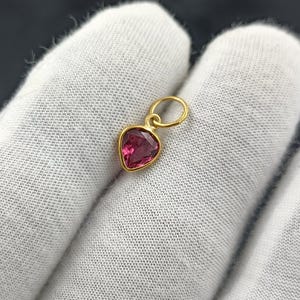 Könnte beinhalten: Ein goldfarbener Anhänger mit einem herzförmigen, tiefrosa Edelstein. Der Anhänger hängt an einer kleinen, kreisförmigen Schlaufe. Der Edelstein ist in eine goldene Lünette gefasst. Der Hintergrund ist ein weißer Stoff.