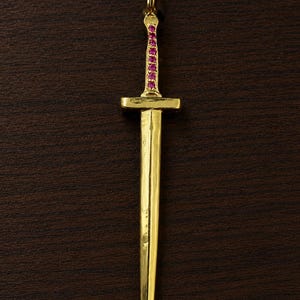 May include: A gold-colored pendant in the shape of a sword. The handle is adorned with a row of small, pink gemstones. The sword has a crossguard and a long, pointed blade. The pendant is suspended from a small loop at the top.