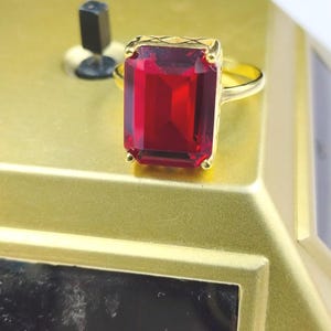 May include: A gold ring with a large, rectangular, deep red gemstone. The ring's setting has a textured design, and the gemstone is the focal point. The ring is set against a gold-coloured surface.