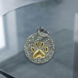 May include: A circular pendant featuring a gold-toned paw print design centered on a silver-toned base covered in small, clear, sparkling stones. The pendant includes a small circular bail at the top for attachment to a chain or necklace.