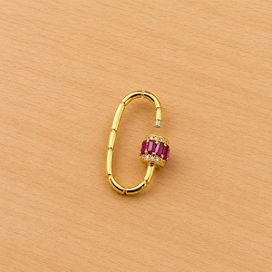 May include: A gold-colored, oval-shaped clasp with a decorative, cylindrical center. The center is adorned with rectangular, ruby-colored stones and small, clear accent stones. The clasp is open, revealing the internal mechanism.