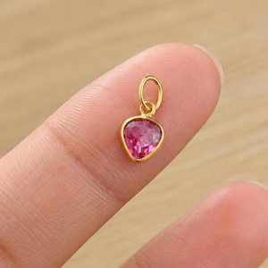 Puede incluir: Un pequeño colgante dorado con una piedra preciosa rosa en forma de lágrima. La piedra está engastada en un bisel dorado y tiene un lazo en la parte superior para sujetarlo a una cadena. El colgante está sujeto por un dedo.