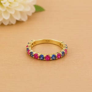 May include: A gold-colored ring with alternating pink and blue gemstones. The ring is set with small, round stones, creating a colorful band. The ring is on a light brown surface.