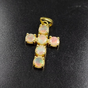 Puede incluir: Un colgante de cruz dorada con ópalos ovalados. Los ópalos presentan una mezcla de tonos blancos, rosas y anaranjados. La cruz se destaca sobre un fondo oscuro, resaltando el diseño y el color del colgante.