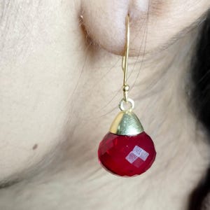 Puede incluir: Un pendiente dorado con una gema roja facetada en forma de lágrima. El pendiente cuelga de un gancho dorado. La gema tiene una tapa dorada.