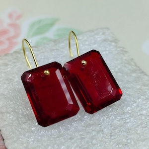 May include: A pair of rectangular, faceted, deep red earrings with gold-colored hooks. The earrings have a small gold accent near the top. The earrings are displayed on a white, textured surface.