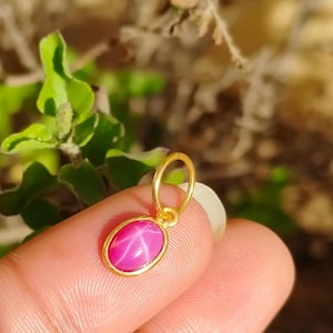 May include: A gold pendant featuring an oval, pink star ruby gemstone. The ruby displays a star pattern in the centre. The pendant is held by a finger against a blurred green and brown backdrop.
