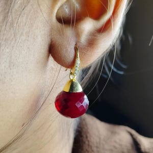 Puede incluir: Primer plano de un pendiente dorado con una cuenta roja facetada en forma de lágrima. El gancho dorado está adornado con pequeñas piedras preciosas transparentes. El pendiente se lleva en la oreja de una persona.