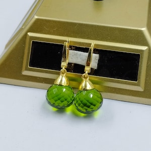 May include: Gold-coloured earrings featuring faceted, olive-green teardrop-shaped gems. The earrings have a lever-back closure and a gold cap connecting the gem to the ear wire. The earrings are set against a gold-coloured background.