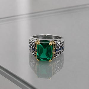 May include: A silver ring with a large, emerald-cut green gemstone. The ring has gold accents around the gemstone and small blue stones on the sides. The band features an ornate design.
