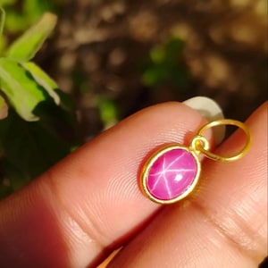 Può includere: Un pendente in oro con una gemma rosa ovale a forma di stella. La gemma è incastonata in una lunetta d'oro e ha un anello d'oro per una catena. Il pendente è tenuto in mano.