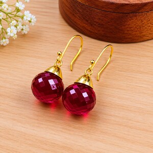 Puede incluir: Un par de pendientes colgantes dorados con colgantes redondos facetados de color rojo rubí. Los pendientes tienen una tapa dorada y un gancho curvo. Los pendientes se exhiben sobre una superficie de madera marrón claro.
