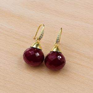 Puede incluir: Un par de pendientes colgantes con gotas de piedras preciosas facetadas de color rojo intenso. Los pendientes tienen detalles metálicos dorados y ganchos para las orejas, con pequeñas piedras transparentes engastadas en la parte superior de los ganchos. Sobre una superficie de madera clara.
