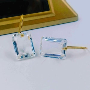 Può includere: Un paio di orecchini rettangolari in cristallo trasparente con ganci color oro. Gli orecchini sono posizionati su uno sfondo bianco, con un elemento decorativo color oro sullo sfondo. I cristalli hanno un taglio sfaccettato.