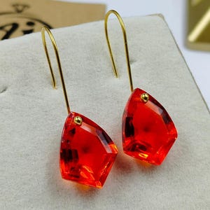May include: A pair of gold-colored drop earrings featuring faceted, vibrant red gemstone pendants. Each pendant has a small gold-colored accent. The earrings hang from delicate gold-colored hooks.