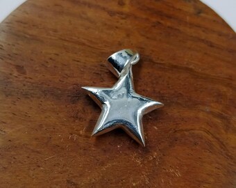 May include: A silver-colored star pendant with a loop for a chain. The five-pointed star is smooth and shiny, reflecting light. The pendant is set against a dark wooden surface.