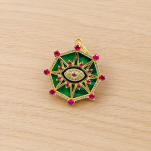 May include: A gold-colored pendant with a green, star-shaped design. The pendant features a central eye design, surrounded by red gemstones and clear crystals. The pendant has a small loop at the top for a chain.