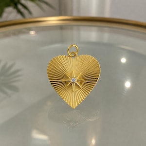 May include: A gold-toned heart-shaped pendant featuring a textured sunburst pattern radiating from the centre. A small, clear, round gemstone is set in the middle of the star-shaped design. The pendant includes a circular jump ring at the top.