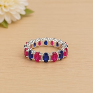 May include: A silver ring featuring a repeating pattern of oval gemstones in alternating shades of pink and blue. The gemstones are set closely together, forming a full circle. The ring is presented on a light wooden surface, with a white flower in the background.