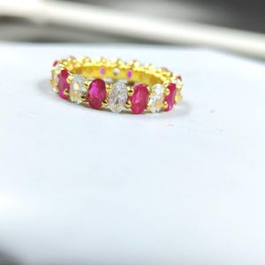 Puede incluir: Un anillo dorado con piedras preciosas rojas y claras de corte ovalado alternadas. El anillo está sobre una superficie blanca, con las piedras preciosas rodeando la banda. El diseño del anillo es simétrico.