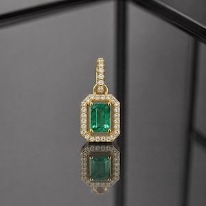 Puede incluir: Colgante de oro con una esmeralda rectangular rodeada de pequeñas piedras preciosas transparentes. El colgante se refleja en la superficie oscura y brillante. El engaste y la argolla también son de oro. La esmeralda es verde brillante.