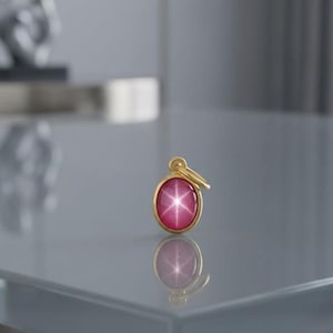 May include: A gold pendant featuring an oval-shaped, deep pink star gemstone. The stone displays a distinct six-rayed star pattern across its surface. The pendant is held by a simple gold bail at the top, reflecting clearly on the smooth surface below.