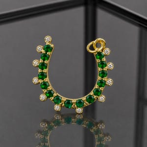 May include: A gold-colored horseshoe-shaped nose ring. The jewelry is adorned with a series of round, emerald-green gemstones and small, clear, round gemstones. The nose ring is displayed against a dark, reflective surface.