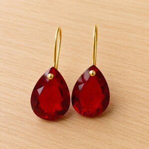 May include: A pair of red gemstone earrings, teardrop-shaped, with gold-coloured hooks and small gold accents. The earrings are presented on a light-coloured wooden surface.
