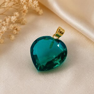 Pode incluir: Um pingente em forma de coração com uma pedra preciosa facetada de cor verde-azulado. O pingente tem uma argola dourada. A pedra preciosa é colocada sobre um fundo cremoso acetinado. Elementos florais decorativos são visíveis.