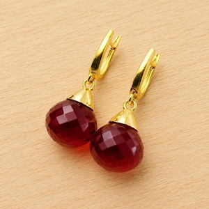 May include: A pair of gold-colored earrings with faceted, deep red gemstone drops. The earrings have a lever back closure and a gold-colored cap that connects the gemstone to the earring hook. The gemstones are a deep, rich red color.