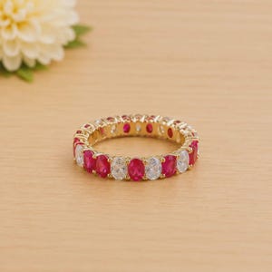 May include: A gold-toned ring with alternating oval-cut red and clear gemstones. The ring is set on a light-colored wooden surface, with a white flower in the background. The ring is a piece of jewelry.