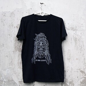 May include: Black t-shirt with a white geometric design of a face and the text "OM NAMAH SHIVAYA!". The t-shirt is hanging on a white hanger against a distressed, off-white wall.