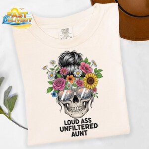May include: Cream-colored t-shirt featuring a skull design with sunglasses and a floral crown. The graphic includes the text "LOUD ASS UNFILTERED AUNT" in black. The shirt has a Comfort Colors tag.
