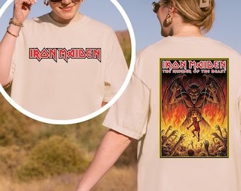 May include: Beige t-shirt featuring the band name "Iron Maiden" in red and black lettering on the front. The back of the shirt displays a graphic from the album "The Number of the Beast" with a devil figure.