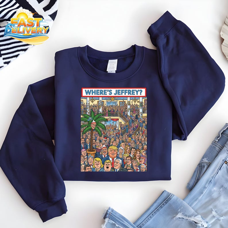 May include: Navy blue sweatshirt featuring a colorful cartoon illustration with the text "WHERE'S JEFFREY?" The graphic depicts a crowd of people with a palm tree and a man's face. The sweatshirt is folded on a white surface.