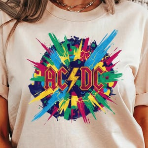 May include: A cream-colored t-shirt featuring a colorful graphic design. The design includes the text "AC/DC" in a bold, maroon font with a yellow lightning bolt, set against a burst of blue, green, yellow, and pink brushstrokes.