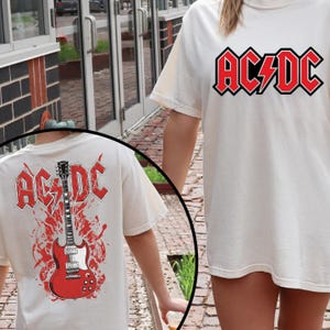 May include: Cream-colored t-shirt featuring the AC/DC logo in red and black. The back of the shirt displays a red electric guitar graphic with the AC/DC logo above it. The shirt is a casual, short-sleeved design.