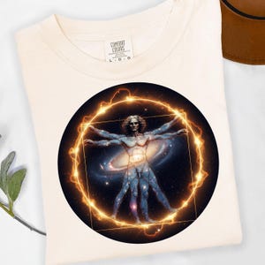 May include: Cream-colored t-shirt featuring a circular graphic of a human figure with arms and legs outstretched, set against a galaxy backdrop. The figure is enclosed within a glowing golden circle and square.