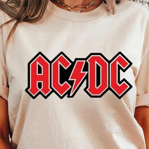 May include: Cream-colored t-shirt featuring the AC/DC logo in red with a black outline. The logo includes the band's name and a lightning bolt graphic. The shirt is made of a soft material and has short sleeves.