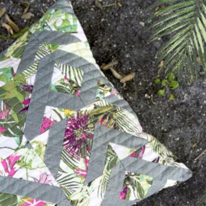 May include: A square throw pillow with a gray and green geometric pattern on a white background. The pillow has a tropical floral print with pink, green, and white flowers.