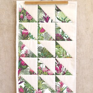 May include: A white quilt with a geometric pattern of green and pink tropical leaf fabric triangles. The quilt is hanging on a wooden hanger.