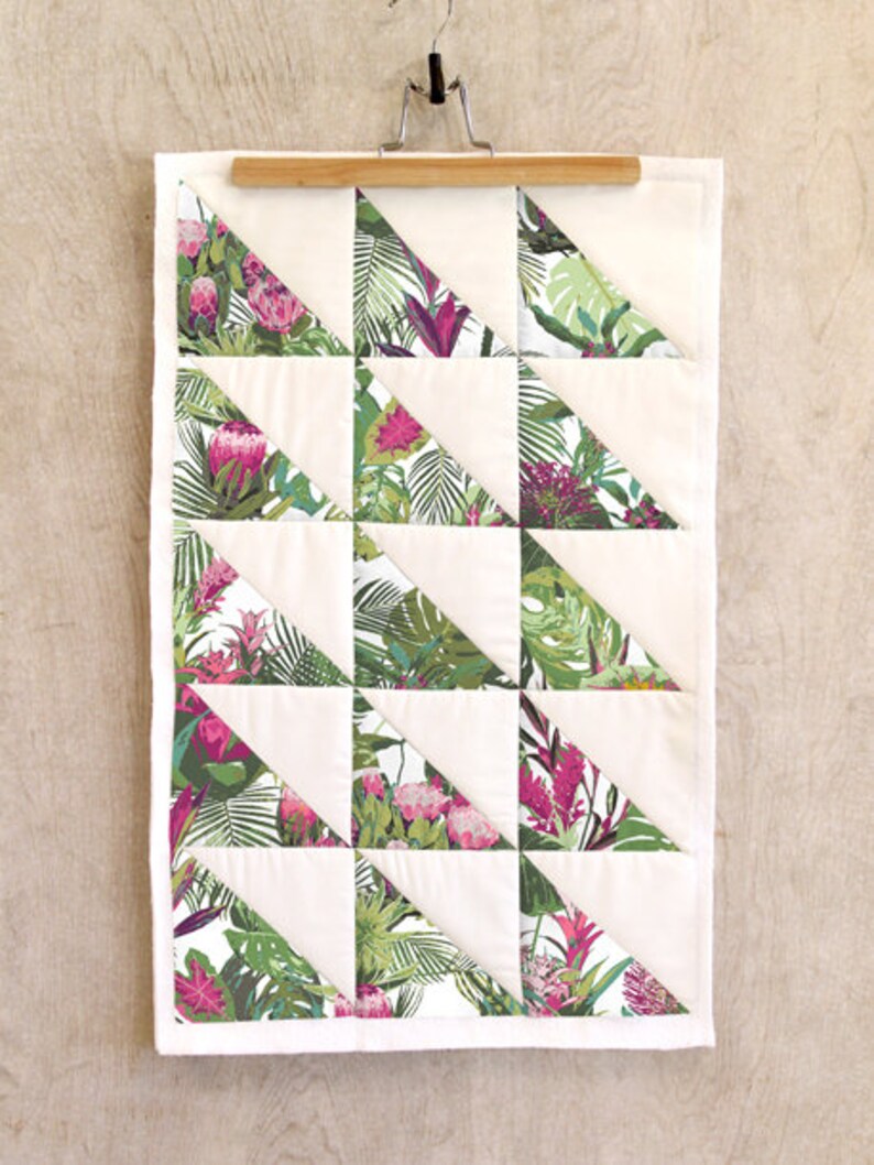 May include: A white quilt with a geometric pattern of green and pink tropical leaf fabric triangles. The quilt is hanging on a wooden hanger.