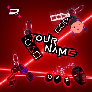Può includere: Portachiavi con vari disegni, tra cui forme, il volto di una ragazza e una maschera, tutti in nero, bianco e rosso. I portachiavi hanno un anello di metallo. È presente anche il testo "YOUR NAME".