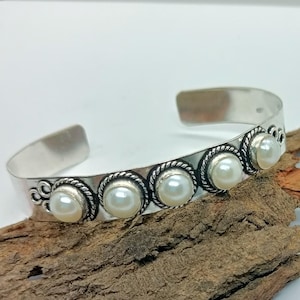 May include: A silver cuff bracelet featuring five round, creamy white pearls set in decorative bezels. The bracelet has an open design and is displayed on a piece of textured wood.